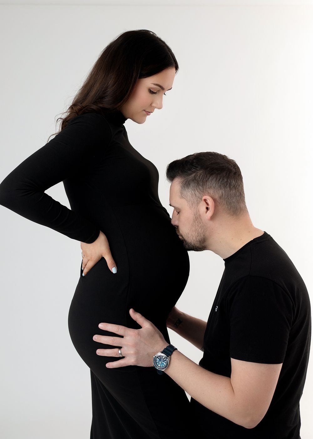 A couple shares intimate moments in black attire against a white backdrop during a maternity photoshoot.