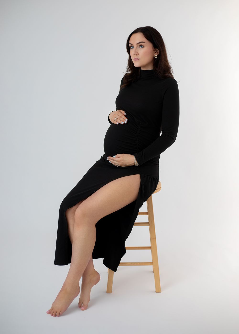 Figure in black sweater dress poses on wooden stool against white backdrop in artistic portrait series.