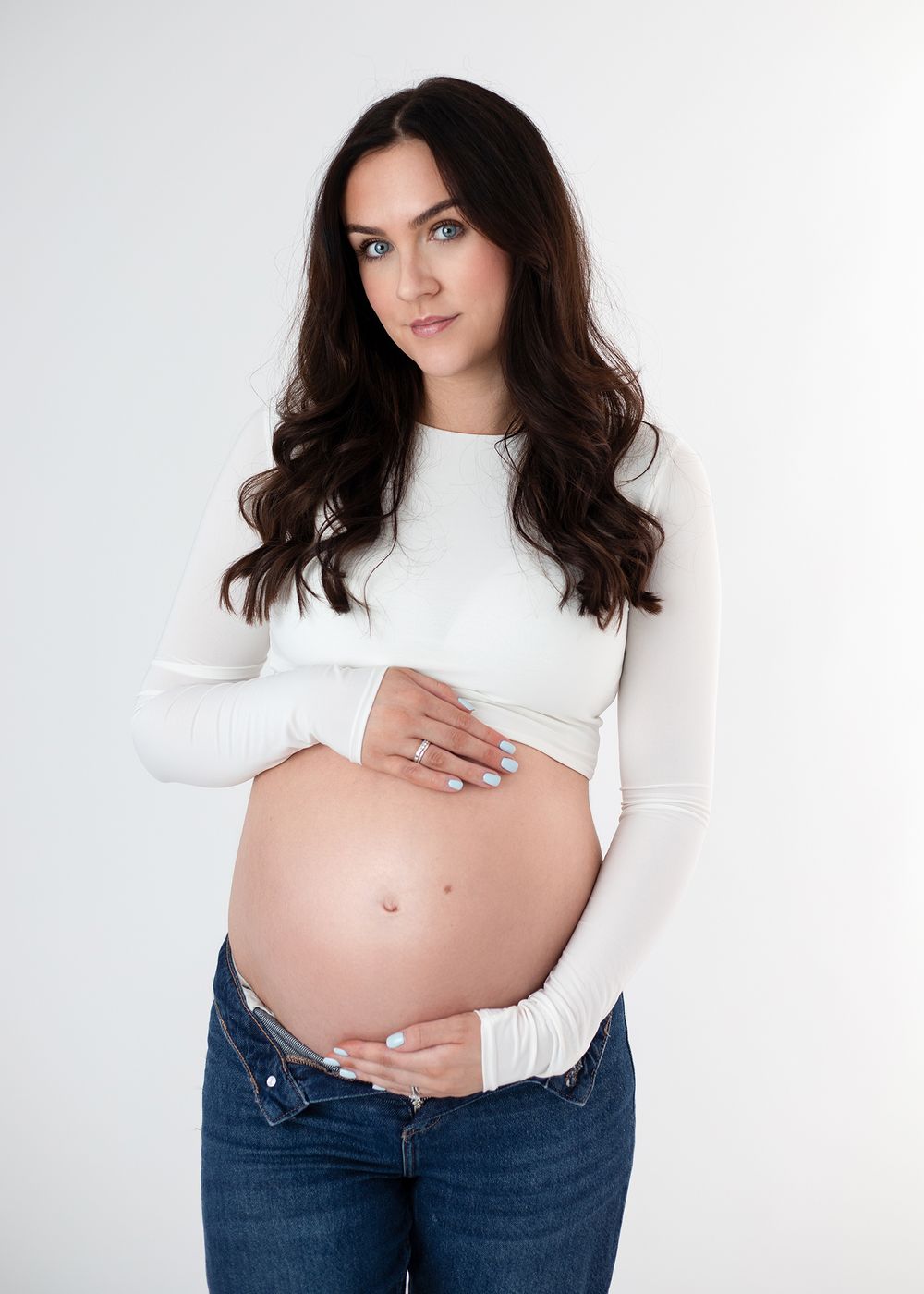 Expectant woman in white long sleeve top and jeans poses against white background while holding her pregnant belly.
