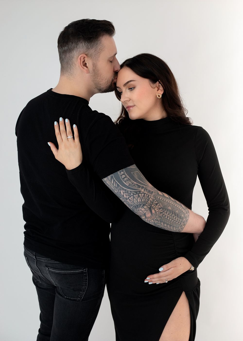 A couple in matching black outfits embraces intimately against a white background, with one person displaying an arm tattoo.