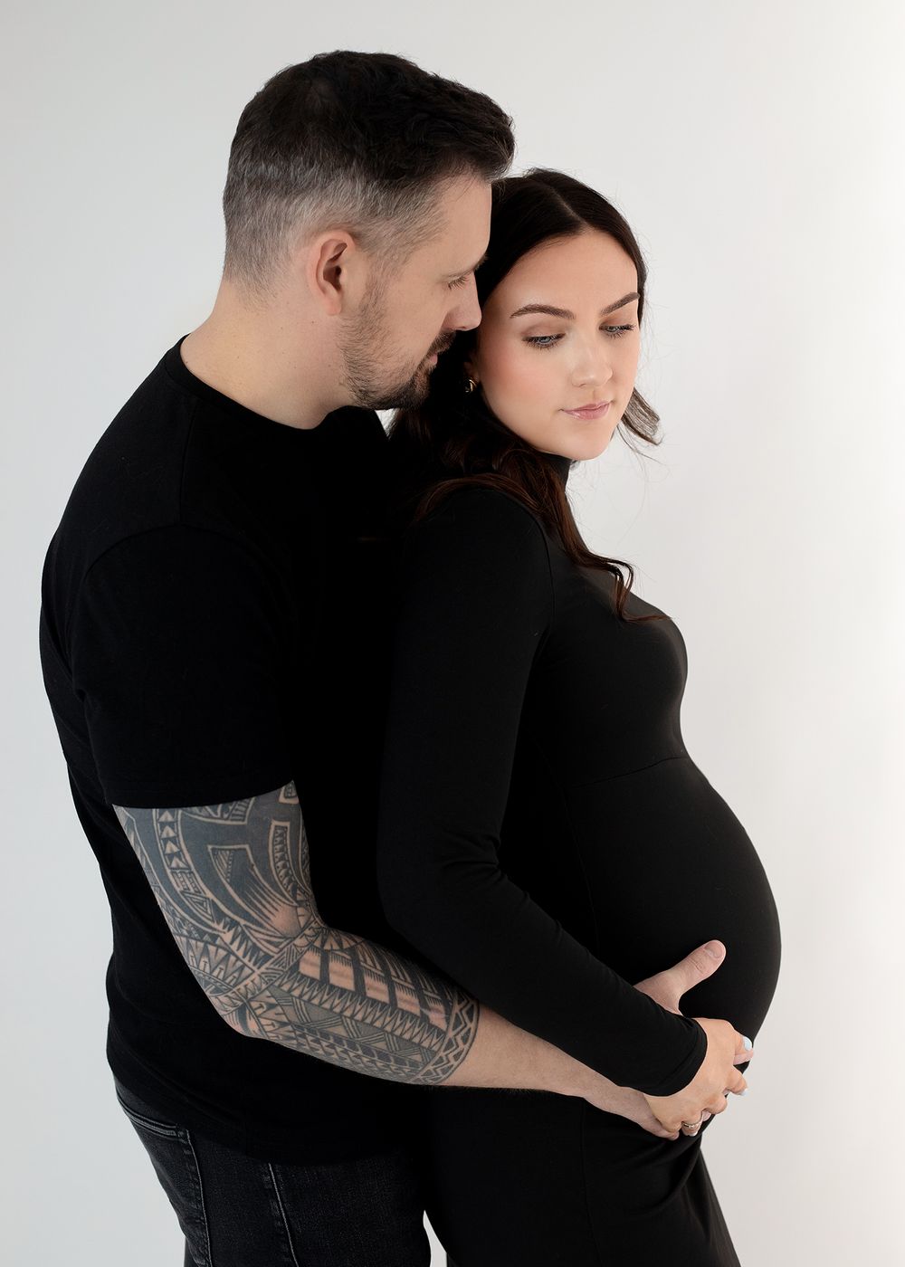 Intimate maternity photo of couple in black clothing embracing against white background, showing tattoo sleeve detail.
