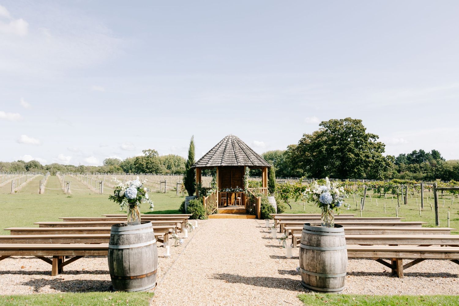 Rustic outdoor wedding venue setup with wooden benches, wine barrels, and a gazebo surrounded by lush greenery.