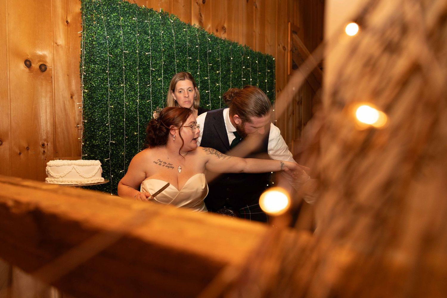 Couple shares an intimate moment in a rustic wooden venue with greenery backdrop and warm lighting.