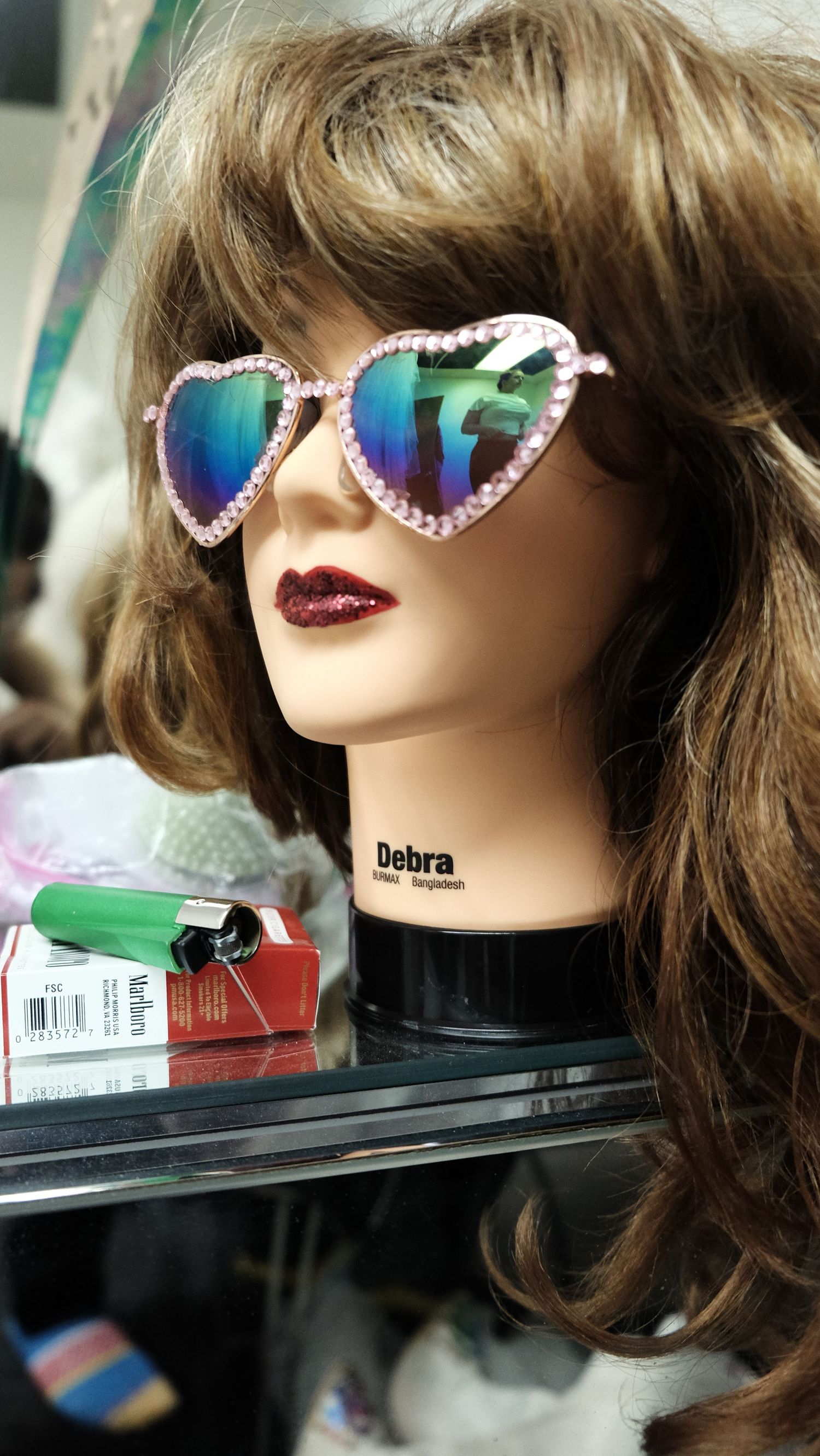 Mannequin head named Debra wearing heart-shaped pink rhinestone sunglasses with rainbow lenses and a brown wig.