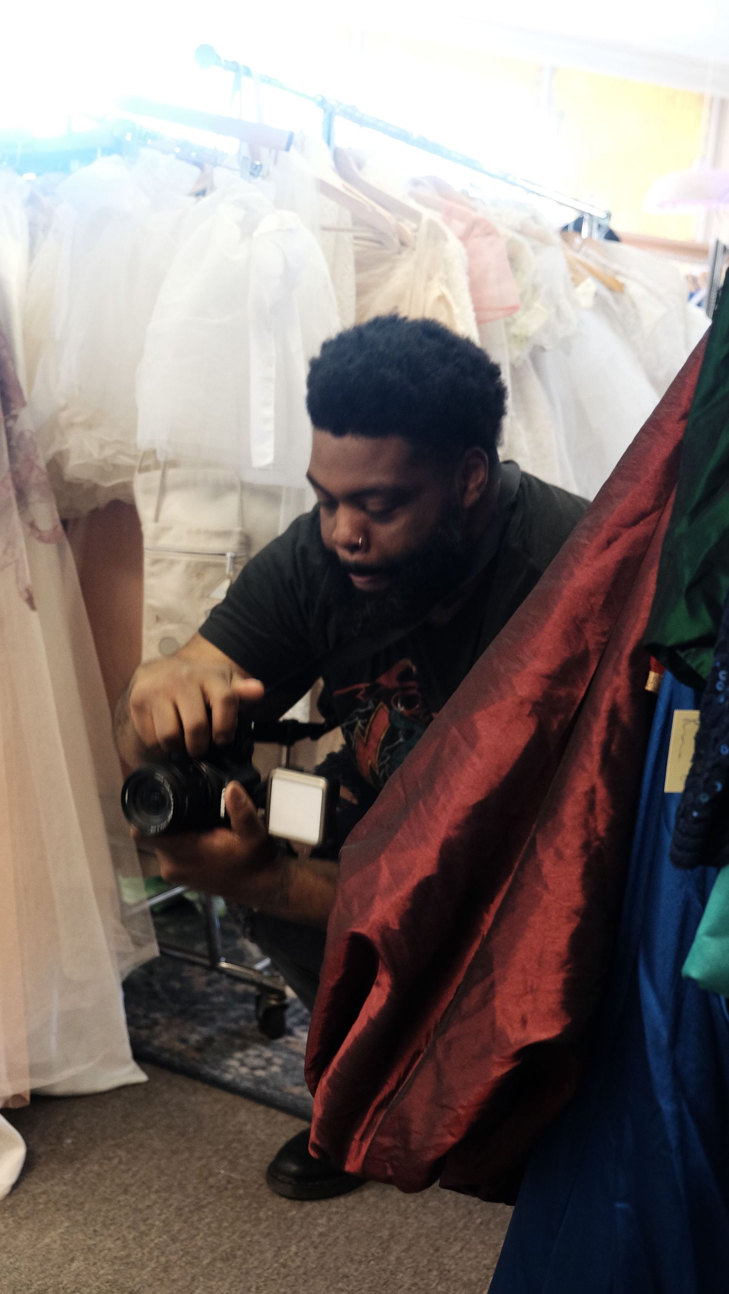 Photographer crouching with camera among wedding dresses on racks in a bridal boutique.