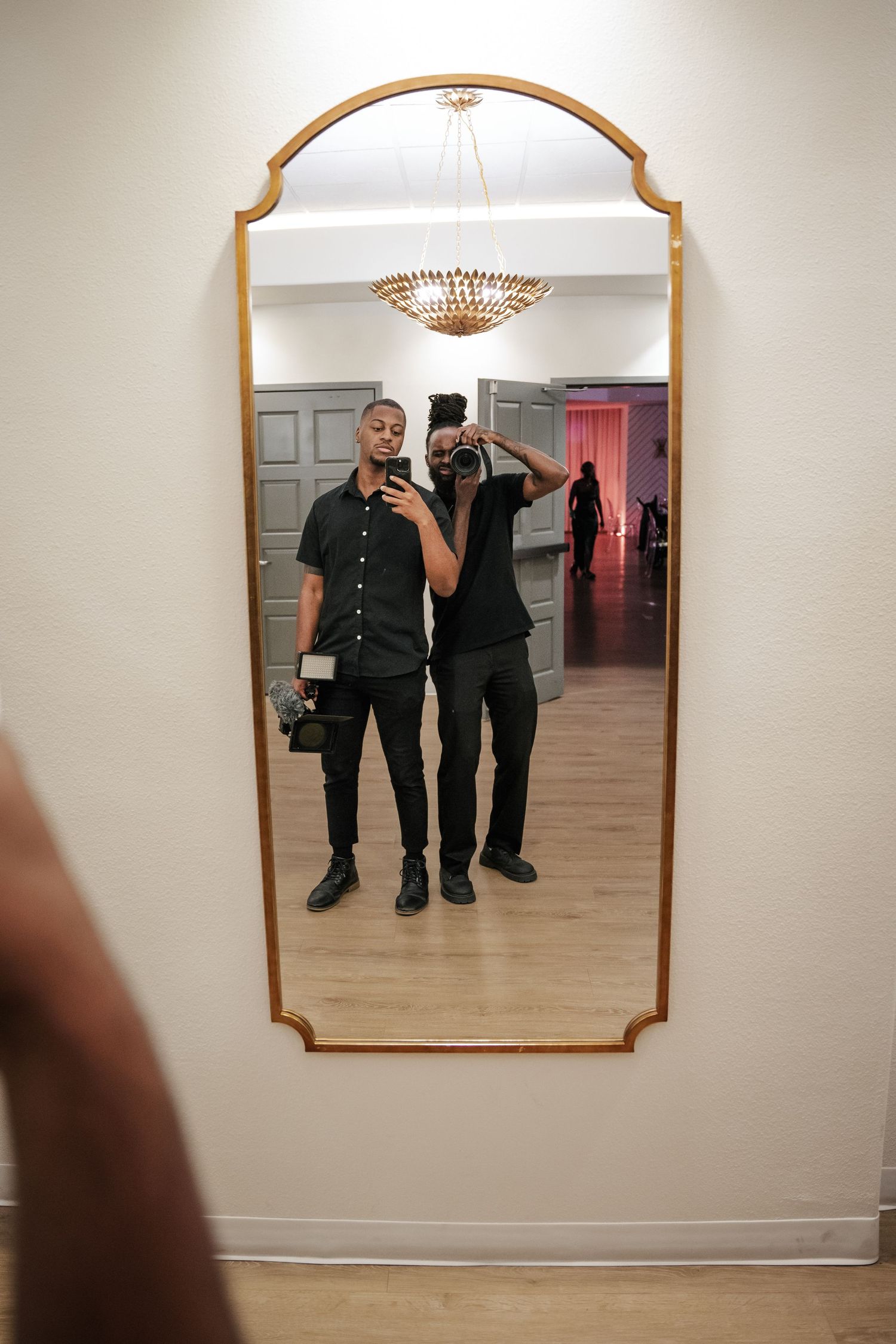 Two people in black outfits taking a mirror selfie in an elegant room with gold-framed mirror and chandelier.