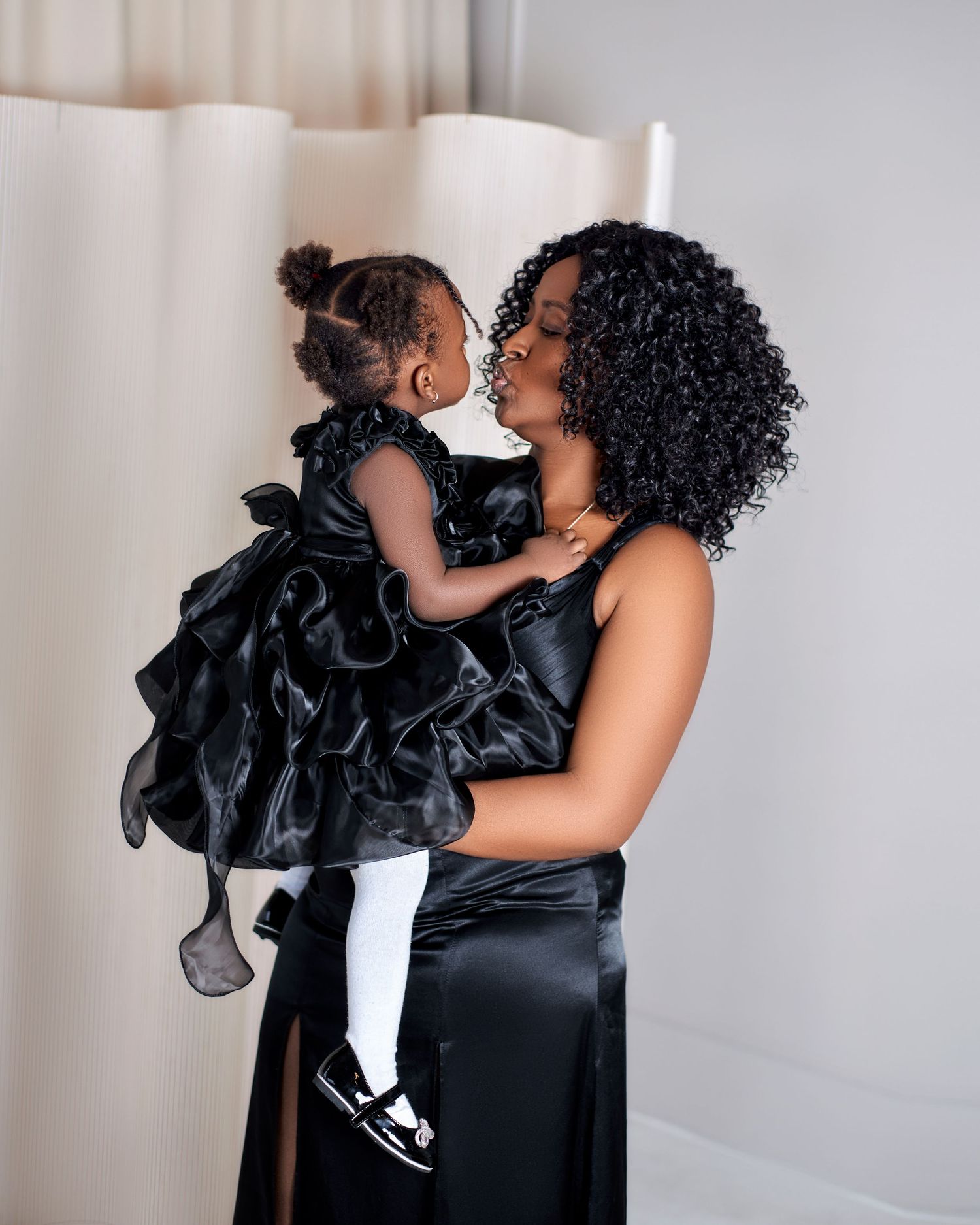 A Black woman in a black satin gown lovingly holds her toddler daughter dressed in a black ruffled dress.