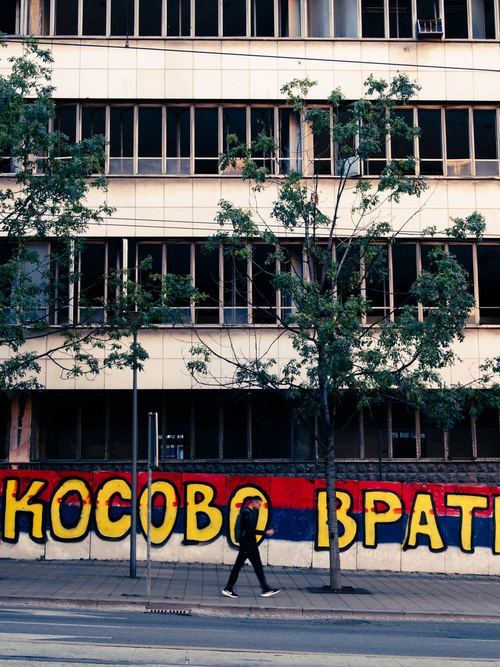 A pedestrian walks past a bold graffiti mural reading KOSOVO VRATI on a weathered city building facade.