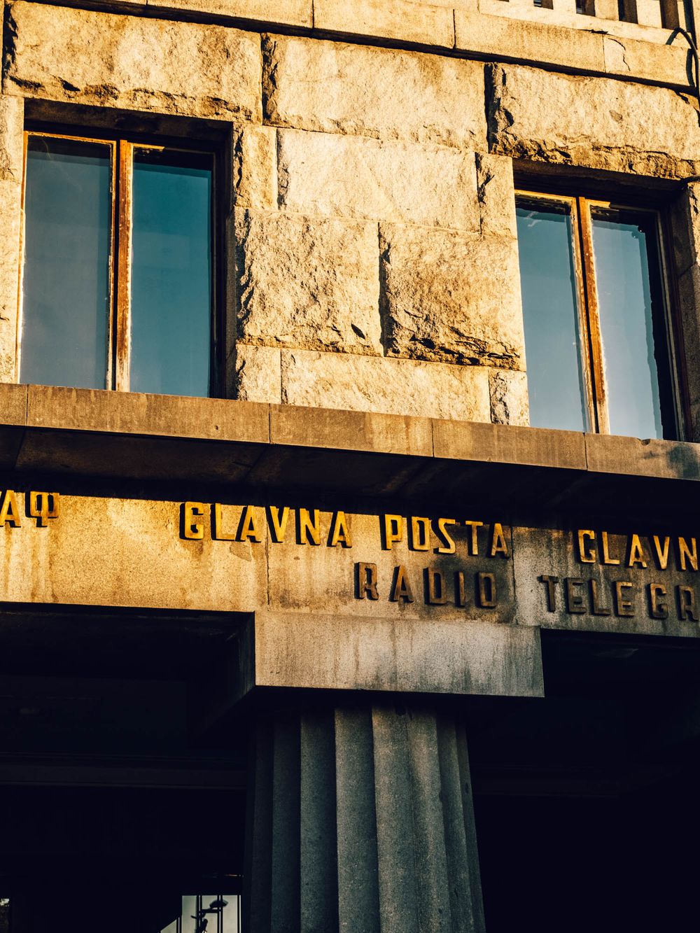 Stone facade of Glavna Posta radio telegraph building with golden lettering and tall windows bathed in warm sunlight.