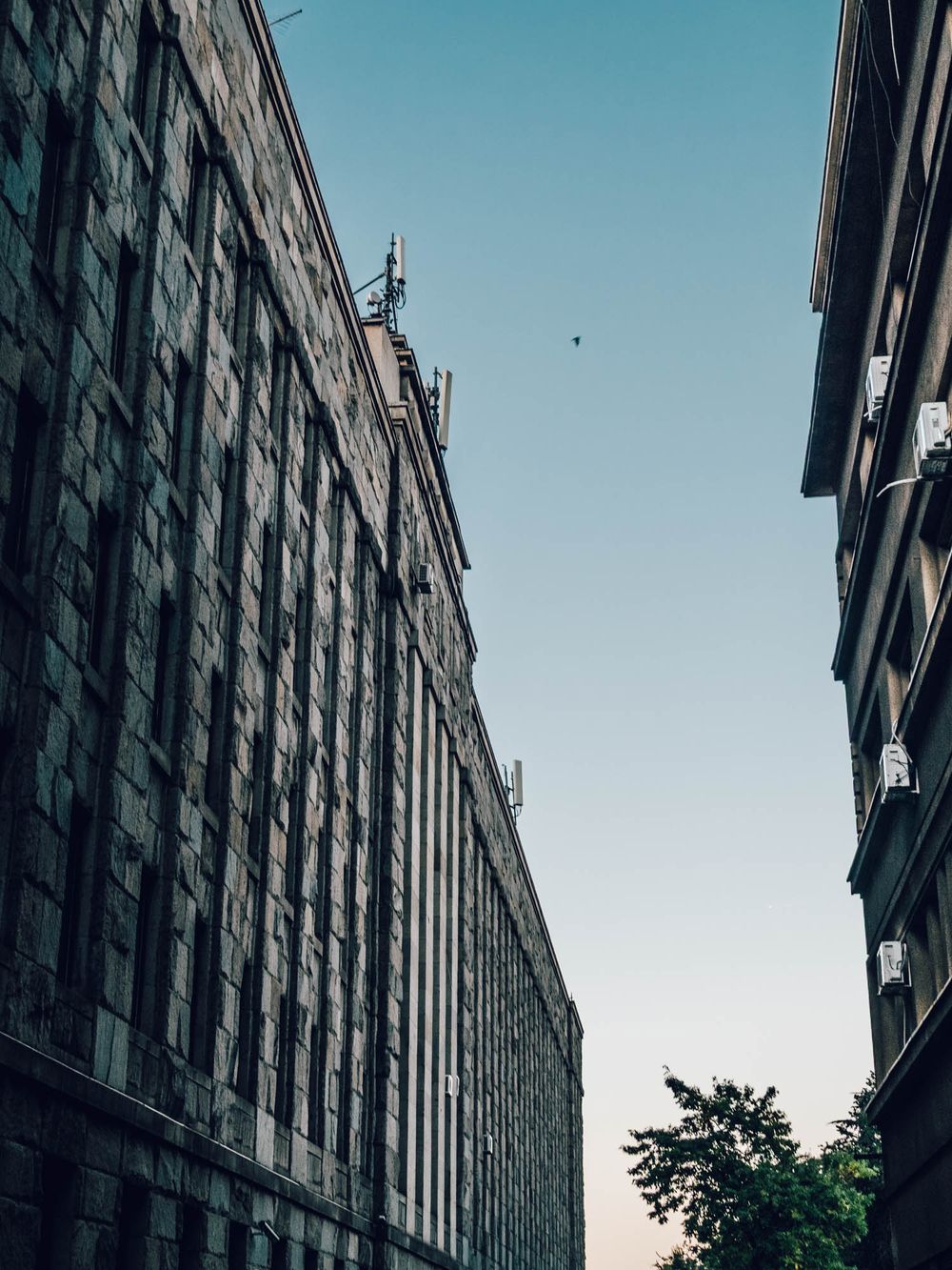 Low-angle view of tall historic stone buildings lining a narrow urban street under a clear blue sky.