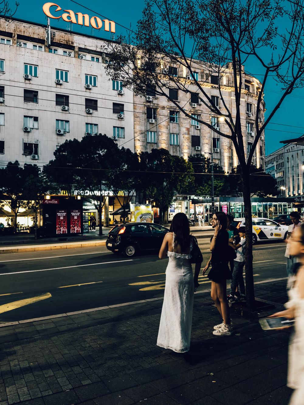 People standing on a city street corner near a Canon building with a McDonald's sign visible at dusk.