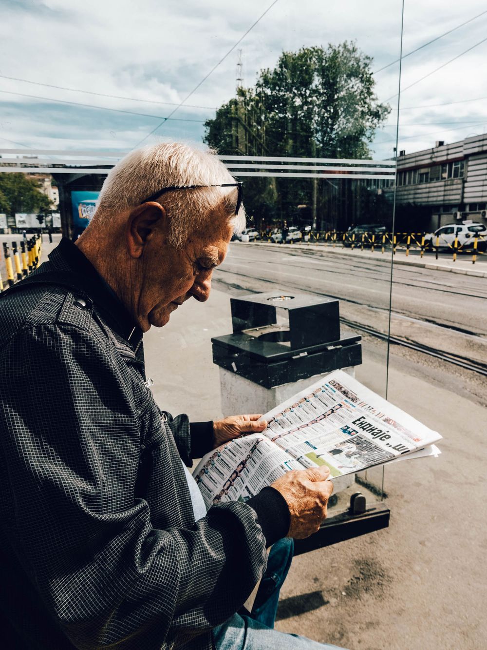 Elderly man reading a newspaper at an outdoor transit stop on a cloudy day.