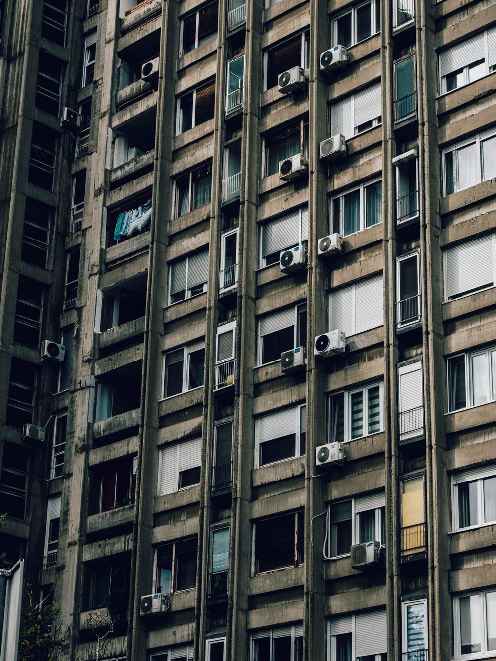 Weathered brutalist apartment building facade with rows of windows and air conditioning units mounted on the exterior walls.