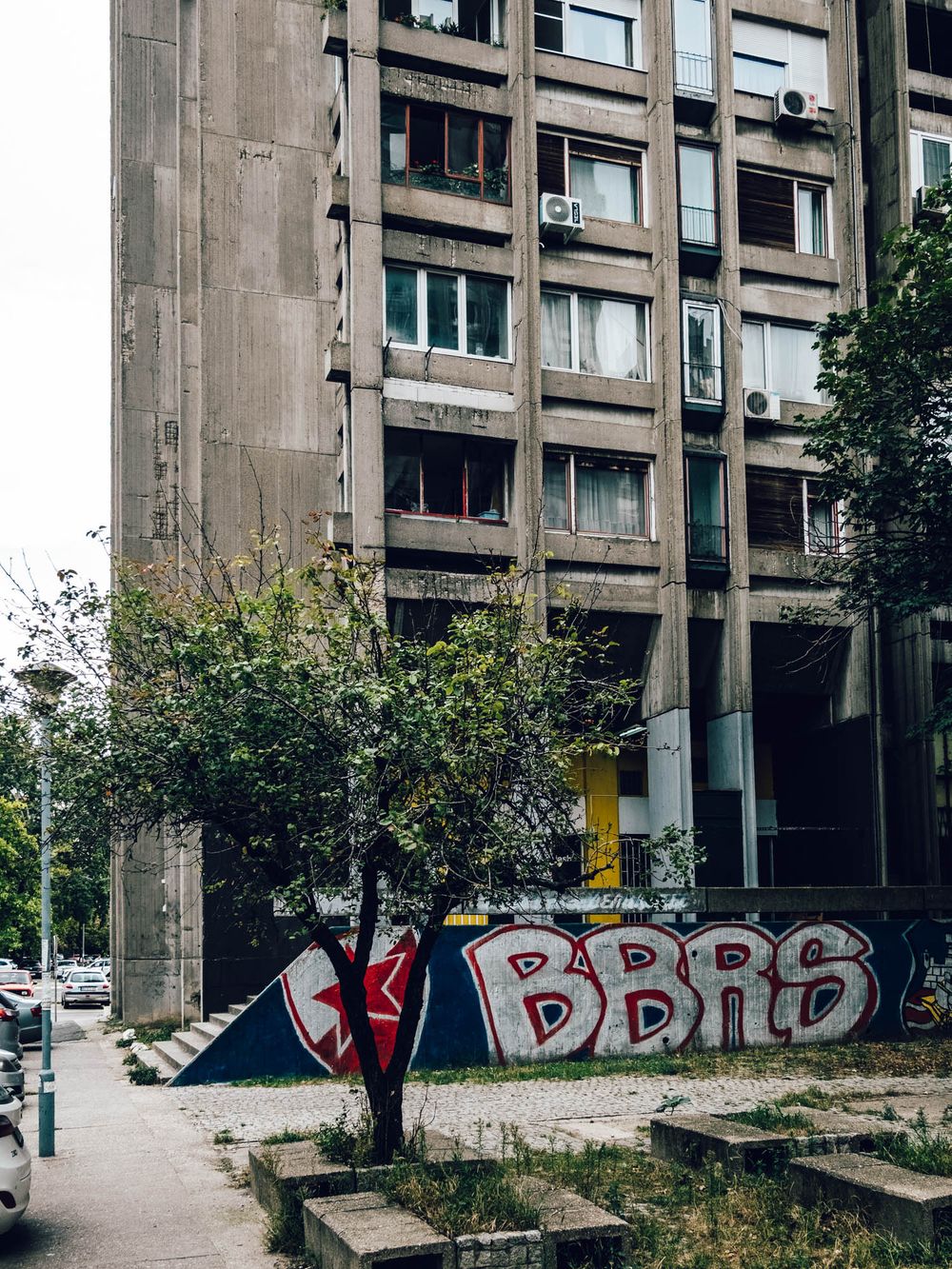 Brutalist apartment building with colorful BBRS graffiti mural on weathered concrete wall, urban street scene.
