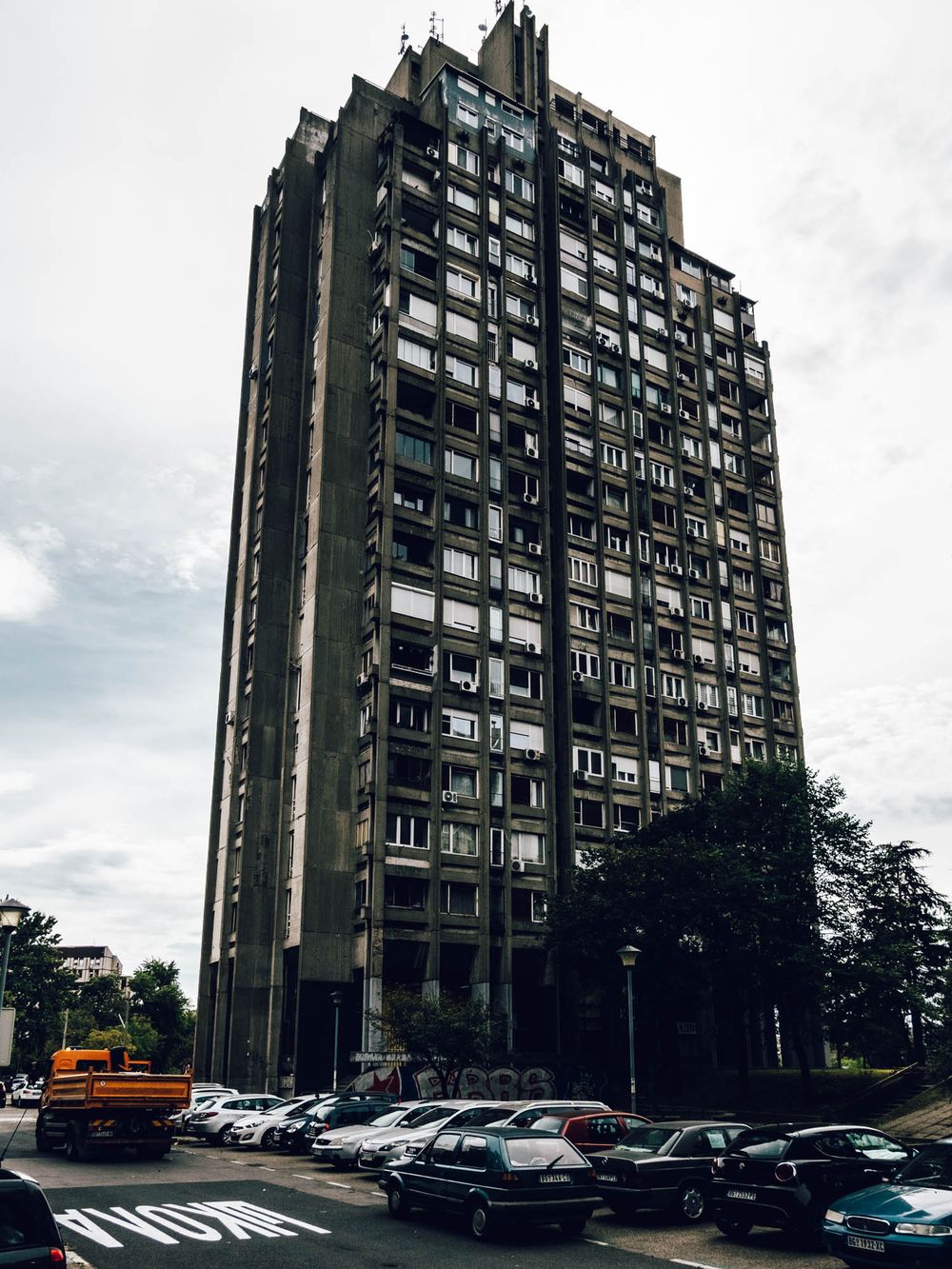 Brutalist high-rise apartment tower with concrete facade, surrounded by parked cars on an overcast day.