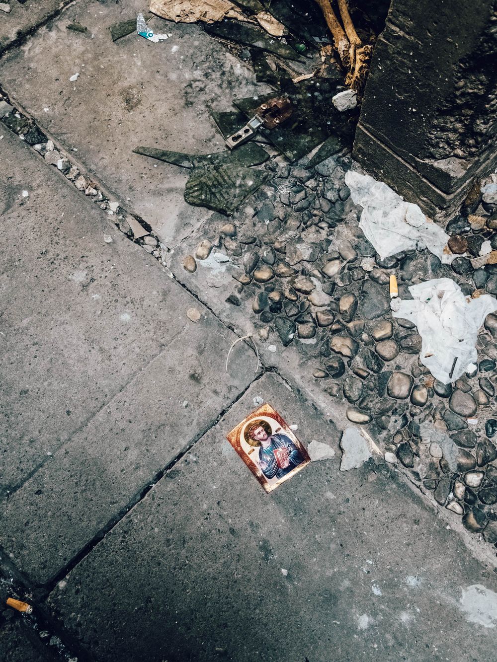 Religious icon card lying on cracked concrete ground surrounded by scattered rocks and debris.