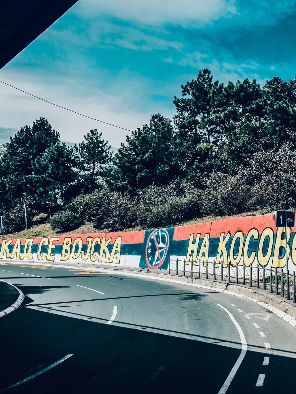 A colorful mural on a retaining wall along a winding road in Serbia, featuring the Serbian flag and Cyrillic text.