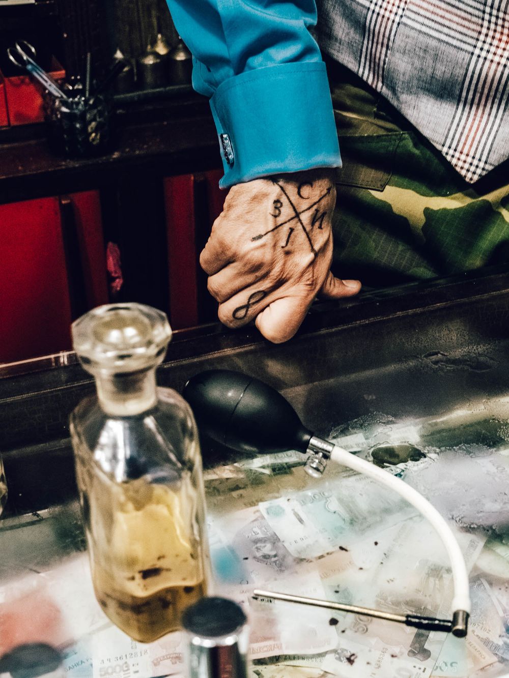 Tattooed hand resting near glass decanter and tubes on a counter, wearing teal jacket.