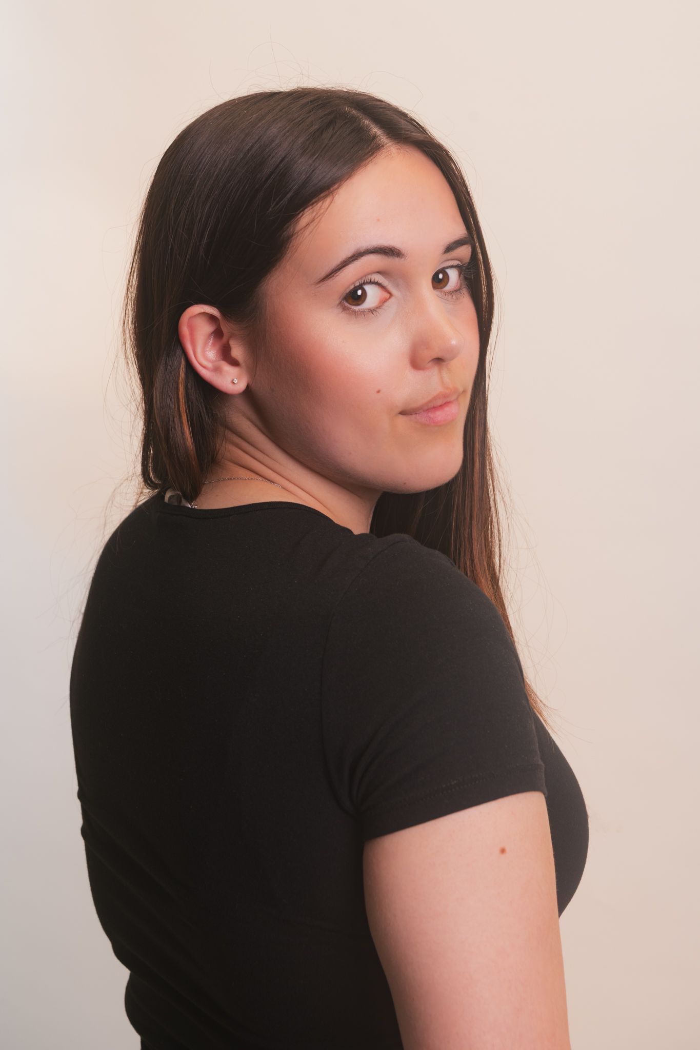 Young woman in black t-shirt glancing over shoulder against neutral background.