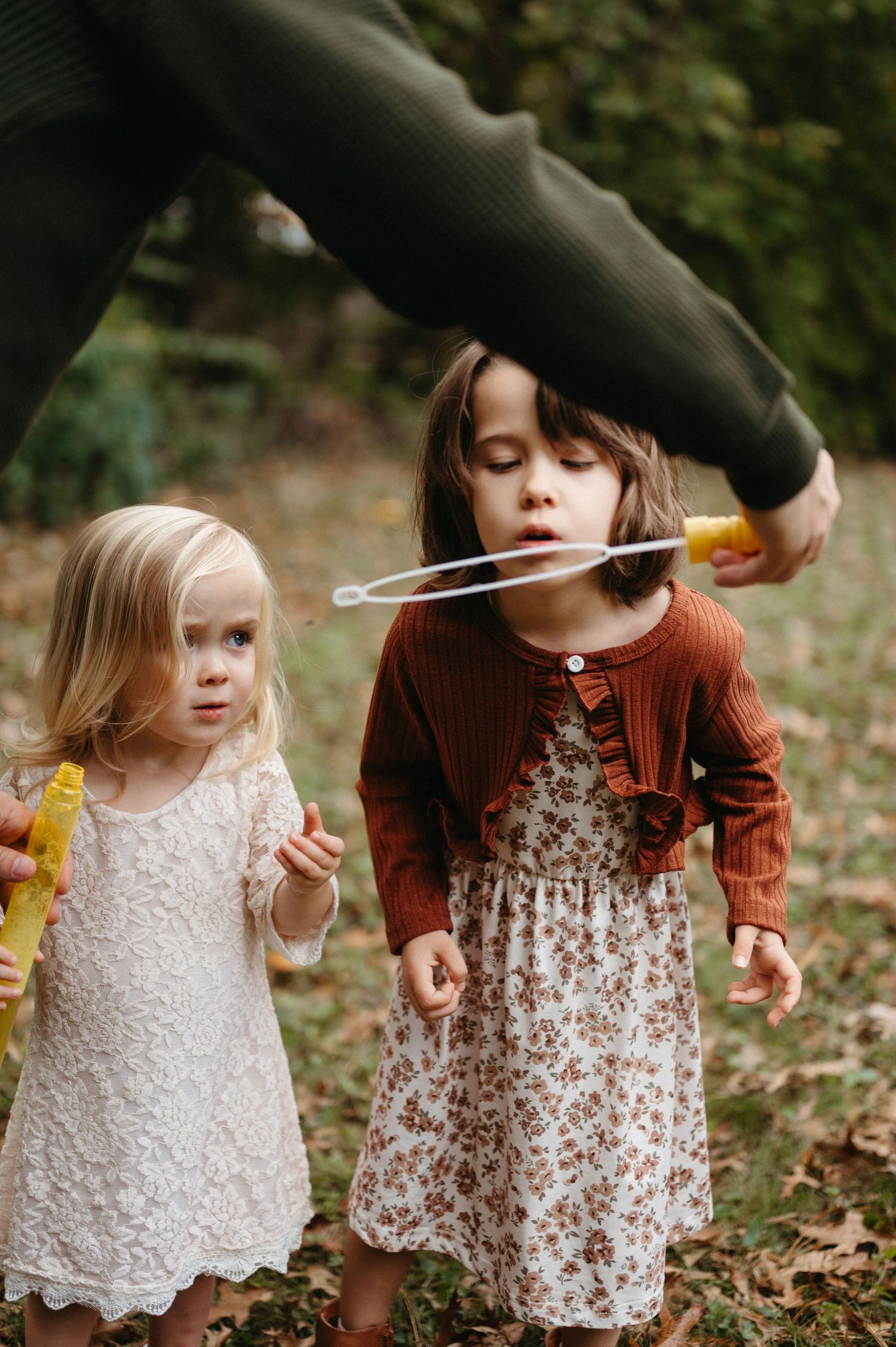 Two young children in dresses sit on autumn leaves while blowing bubbles together in an outdoor setting.