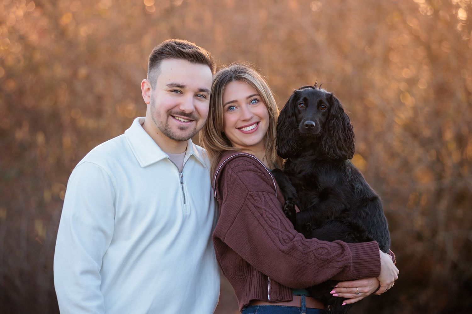 A couple poses outdoors with their black dog during a fall photoshoot with warm autumn colors in the background.
