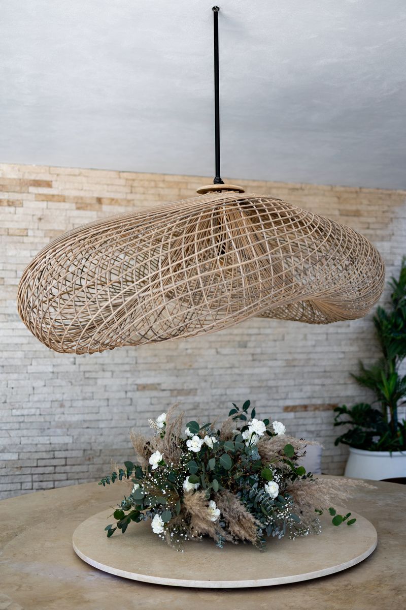 A woven rattan pendant lamp hangs over a rustic floral centerpiece on a round table against a white brick wall.
