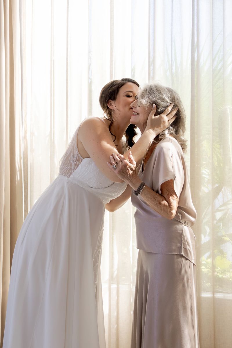 A tender moment shared between a bride in a white gown and an older woman in beige beside a sunlit window.