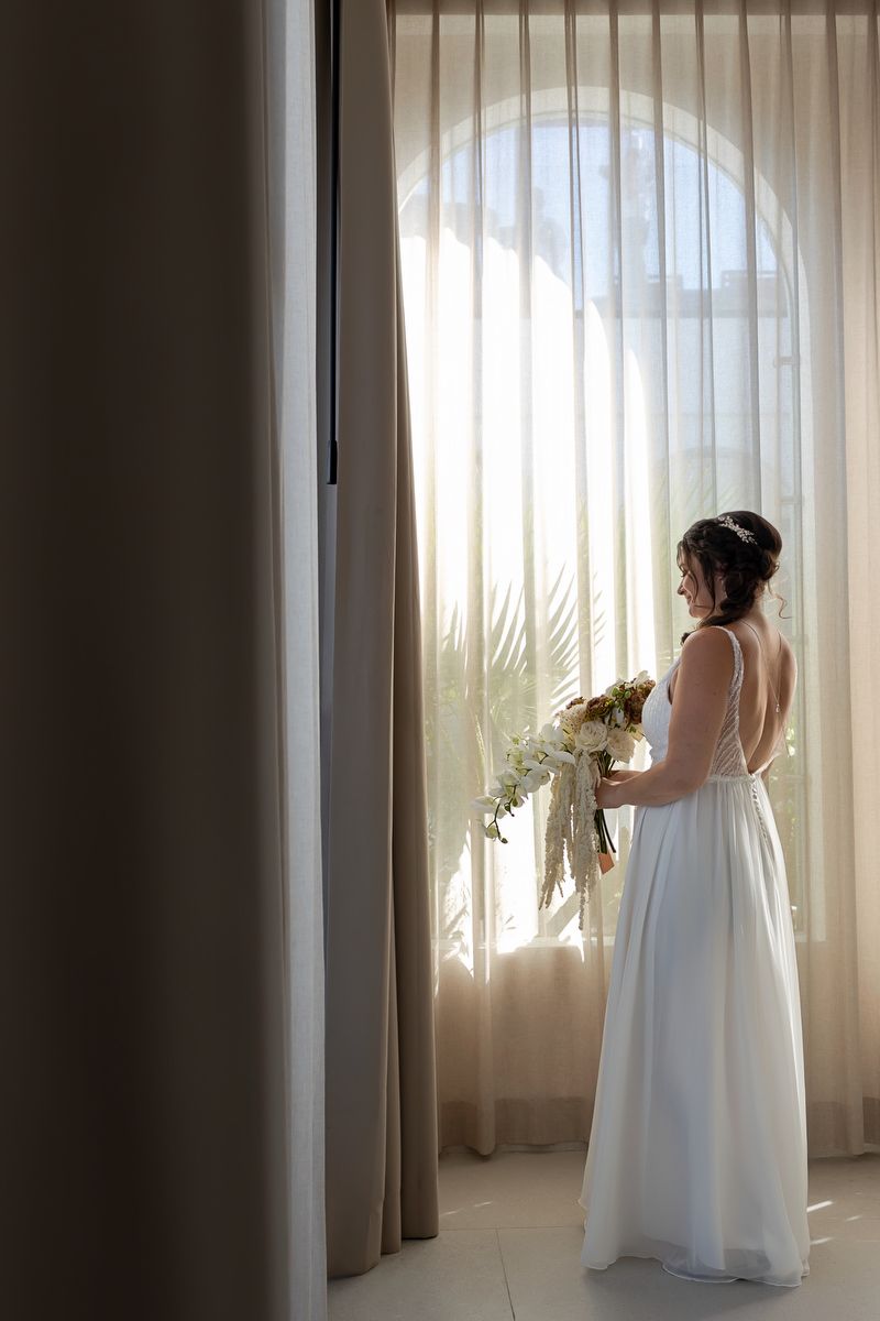 A bride in a white wedding dress stands near sheer curtains in a sunlit room, creating a dreamy silhouette.