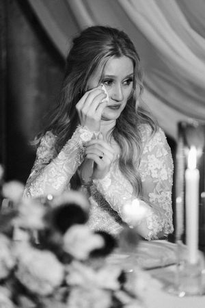 A bride in a long-sleeved lace wedding dress wipes away tears during an emotional moment at her reception.