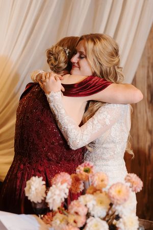 Two people share an emotional embrace at a wedding celebration with pink floral arrangements and a cream backdrop.