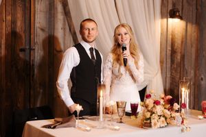 Couple gives wedding reception speeches at elegant rustic venue with floral centerpiece and candlelight on table.