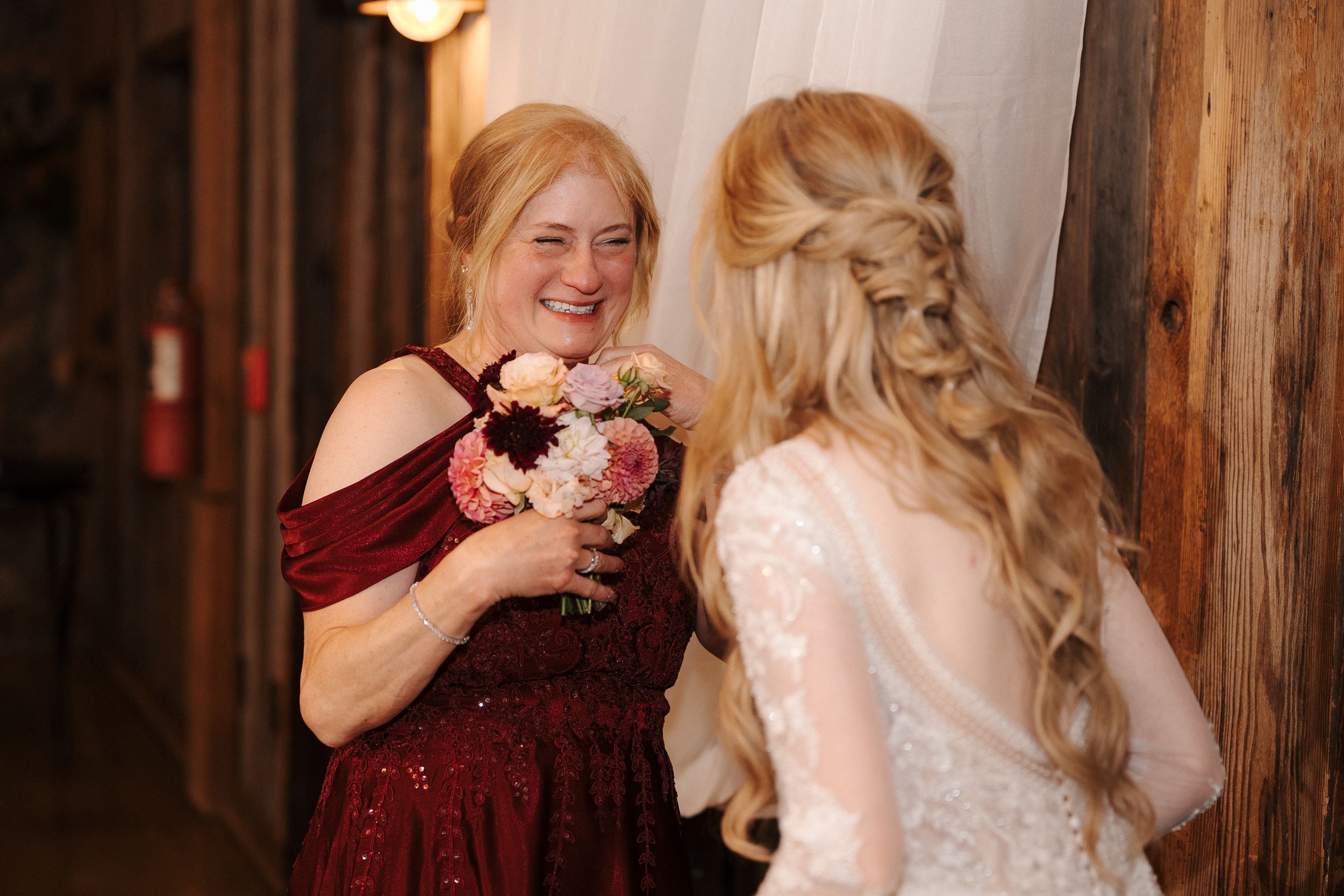 A bride in a white dress shares a joyful moment with someone wearing burgundy at an indoor venue.