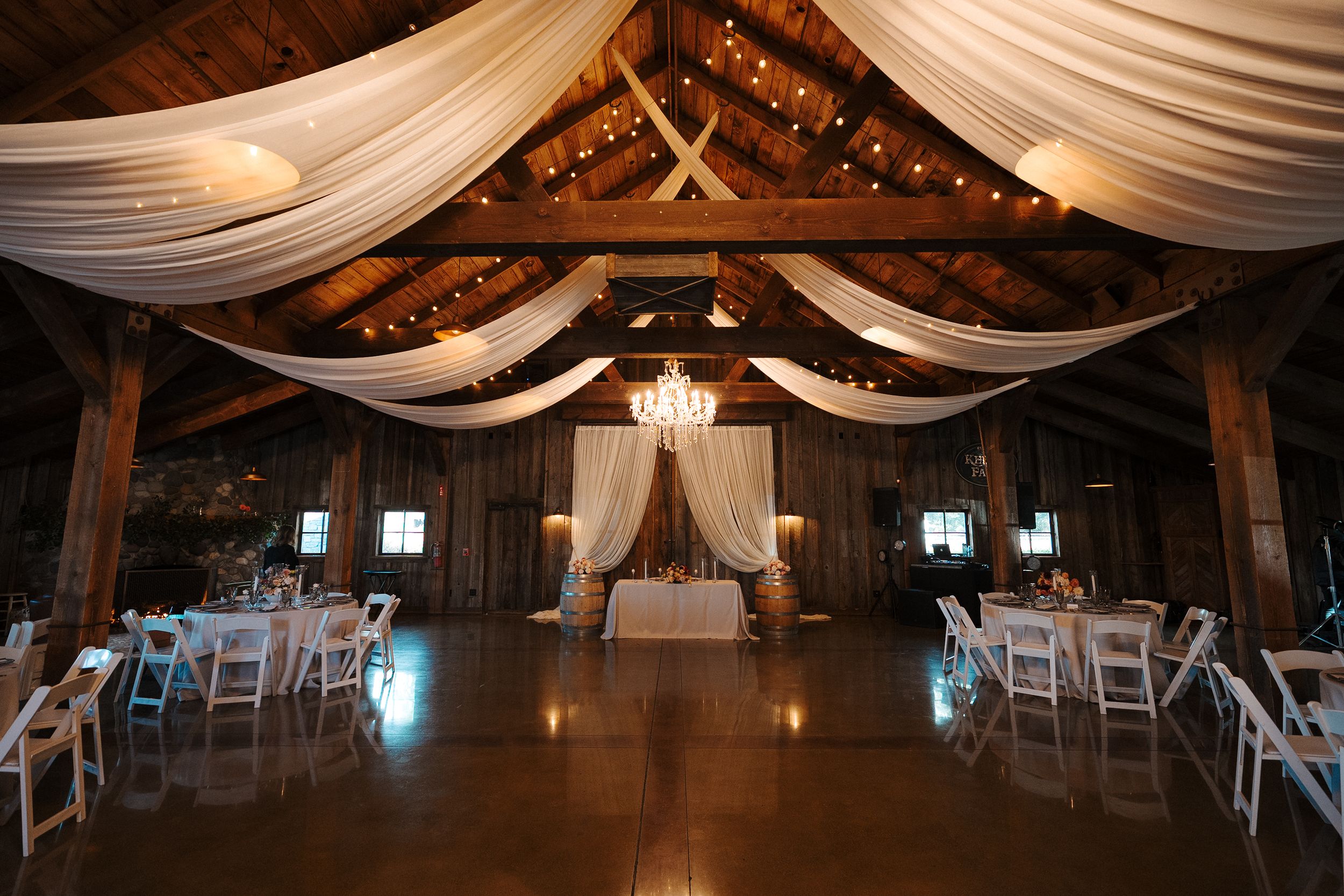 Rustic barn wedding venue interior decorated with elegant white fabric draping and crystal chandeliers for a romantic ceremony.