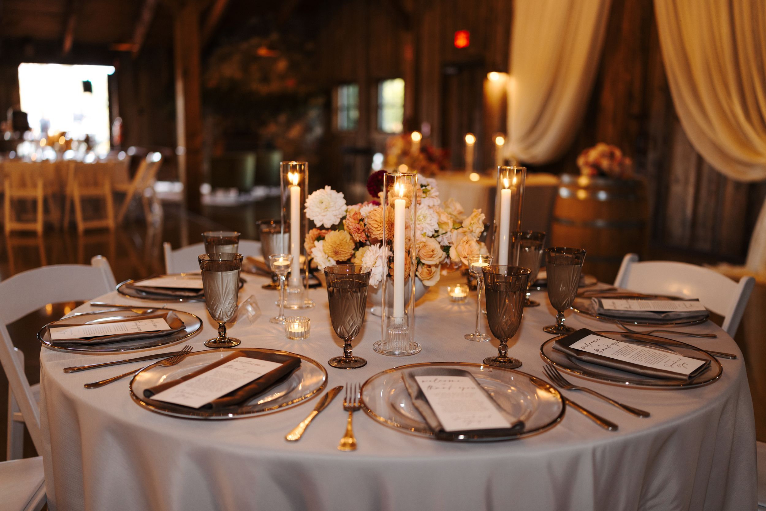 Round dining tables set with white linens and place settings in a rustic barn reception space with ambient lighting.