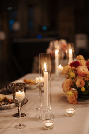 Elegant wedding reception table setting with glowing candles in glass holders and floral centerpieces on white tablecloth.