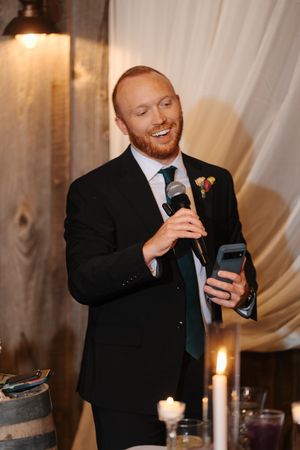 Wedding guest in black suit smiles while reading from phone during reception speech with candlelit decor.