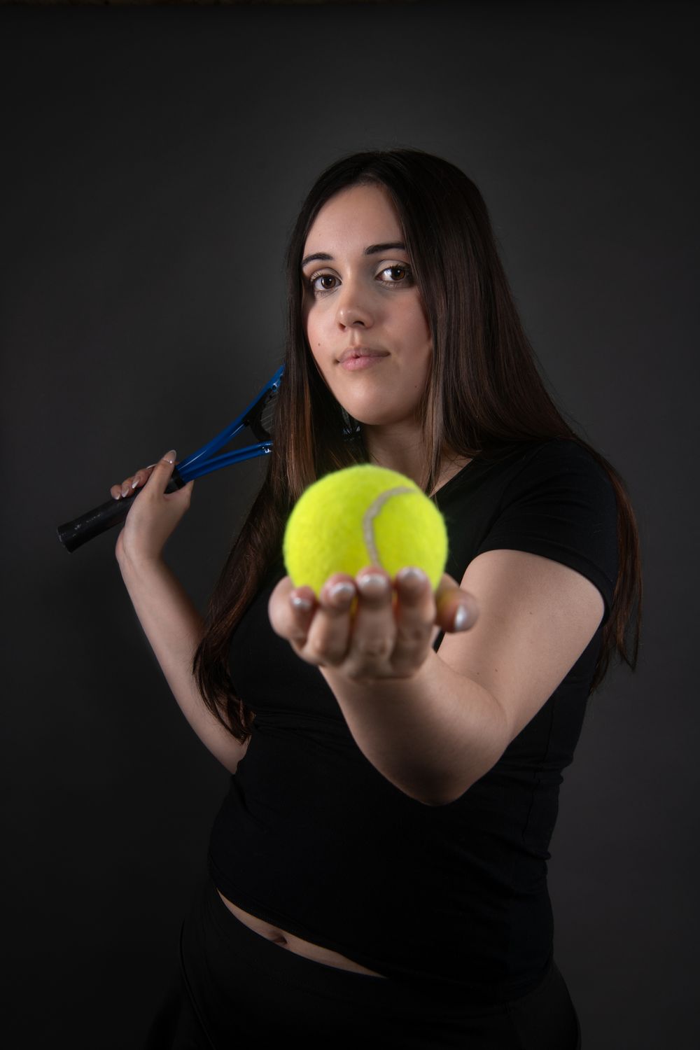 Young woman in black outfit holding tennis racket and extending tennis ball toward camera.