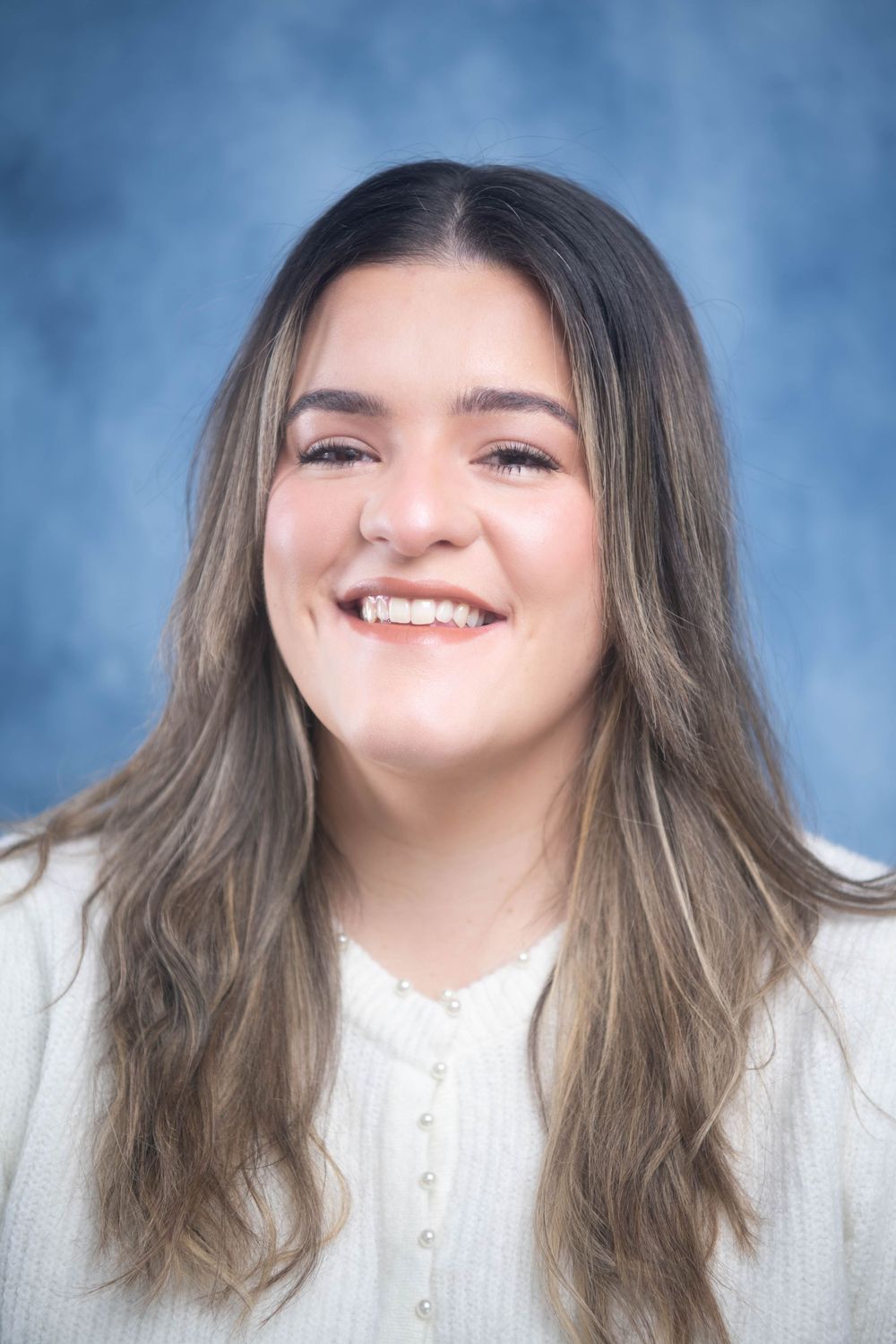 Smiling young woman with long highlighted hair wearing white cardigan against blue background.