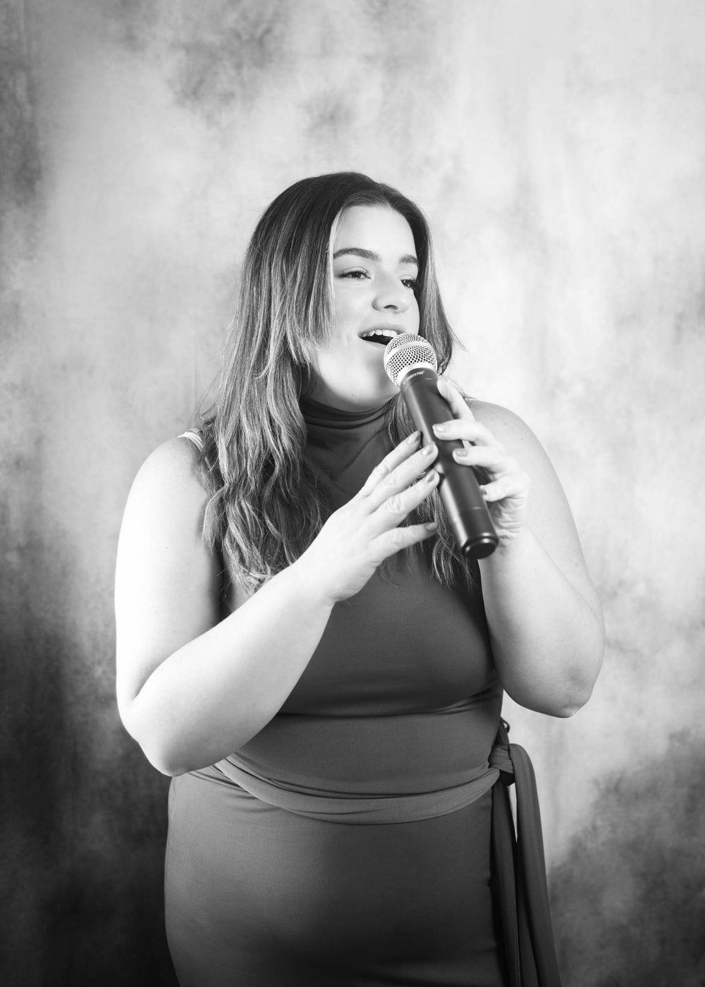 Black and white portrait of a young woman singing into a microphone against a dramatic smoky background.