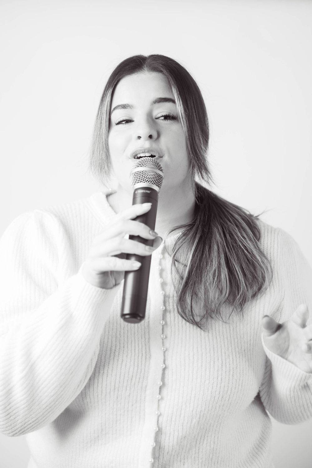 Black and white photo of young woman singing into microphone wearing white knit cardigan.