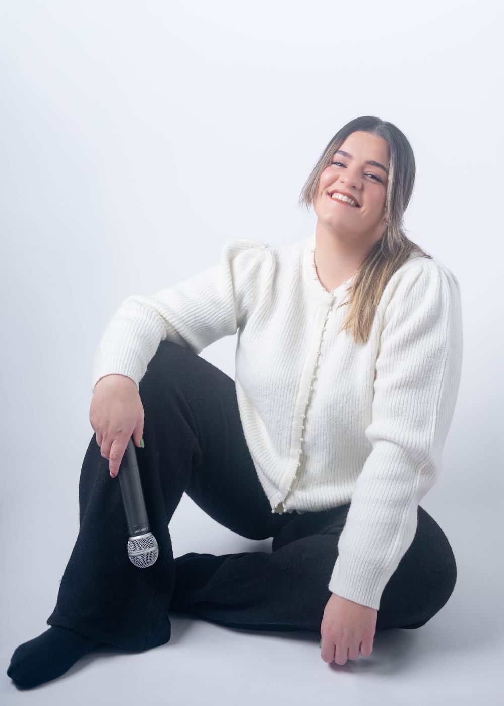 Young woman in white cardigan and black pants sitting on floor smiling while holding microphone.
