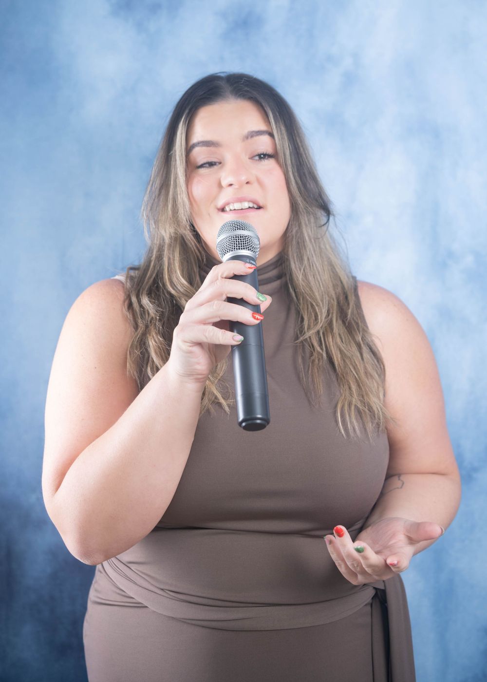 Young woman in brown sleeveless dress singing passionately into microphone against blue backdrop.