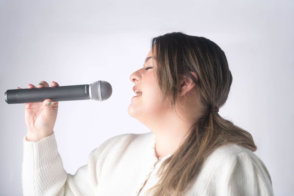 Young woman singing passionately into a wireless microphone wearing a white knit sweater against a grey background.