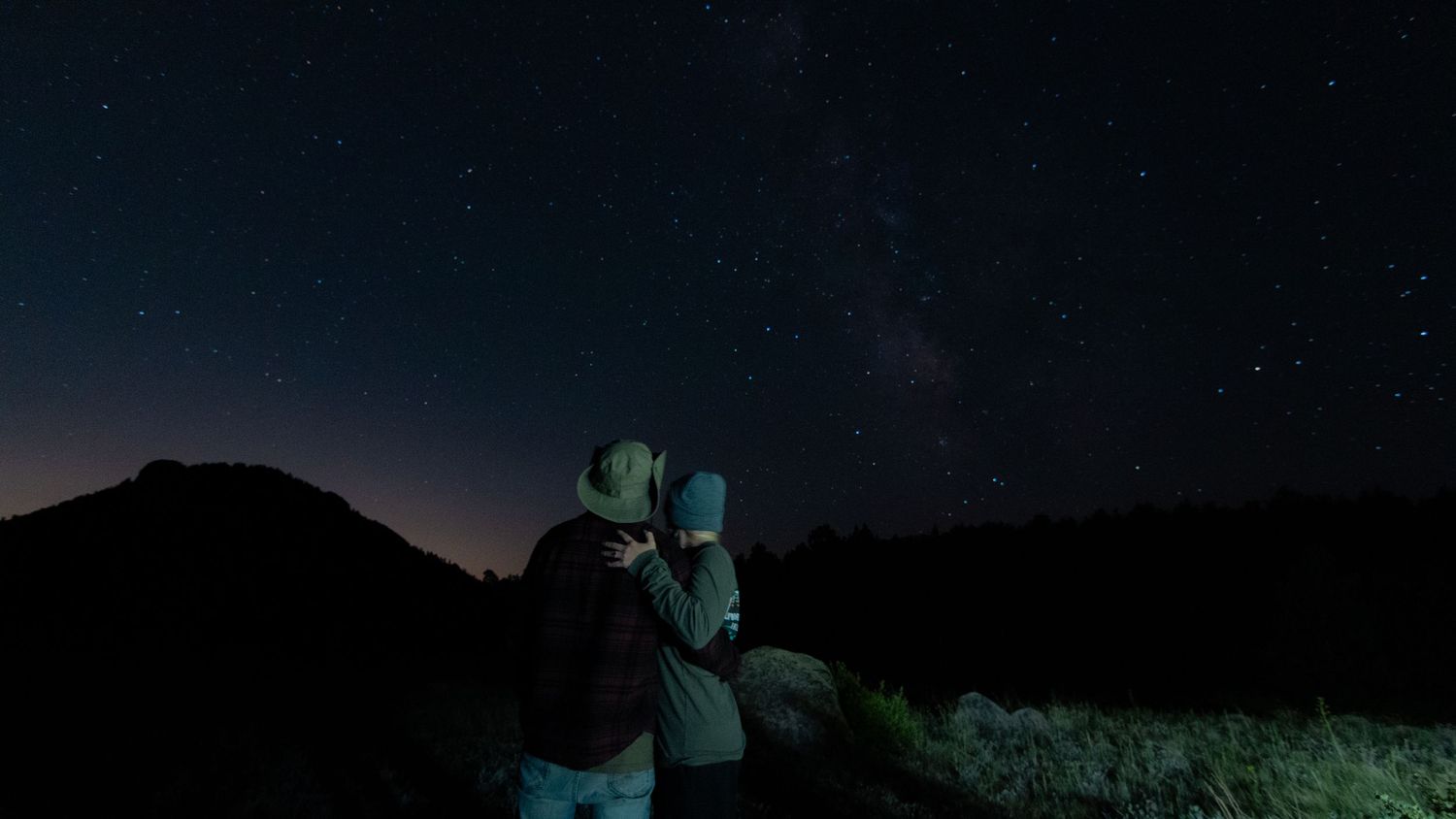 A starry night sky stretches over dark a romantic couple with rolling hills in a panoramic view in Rocky Mountain National Park CO