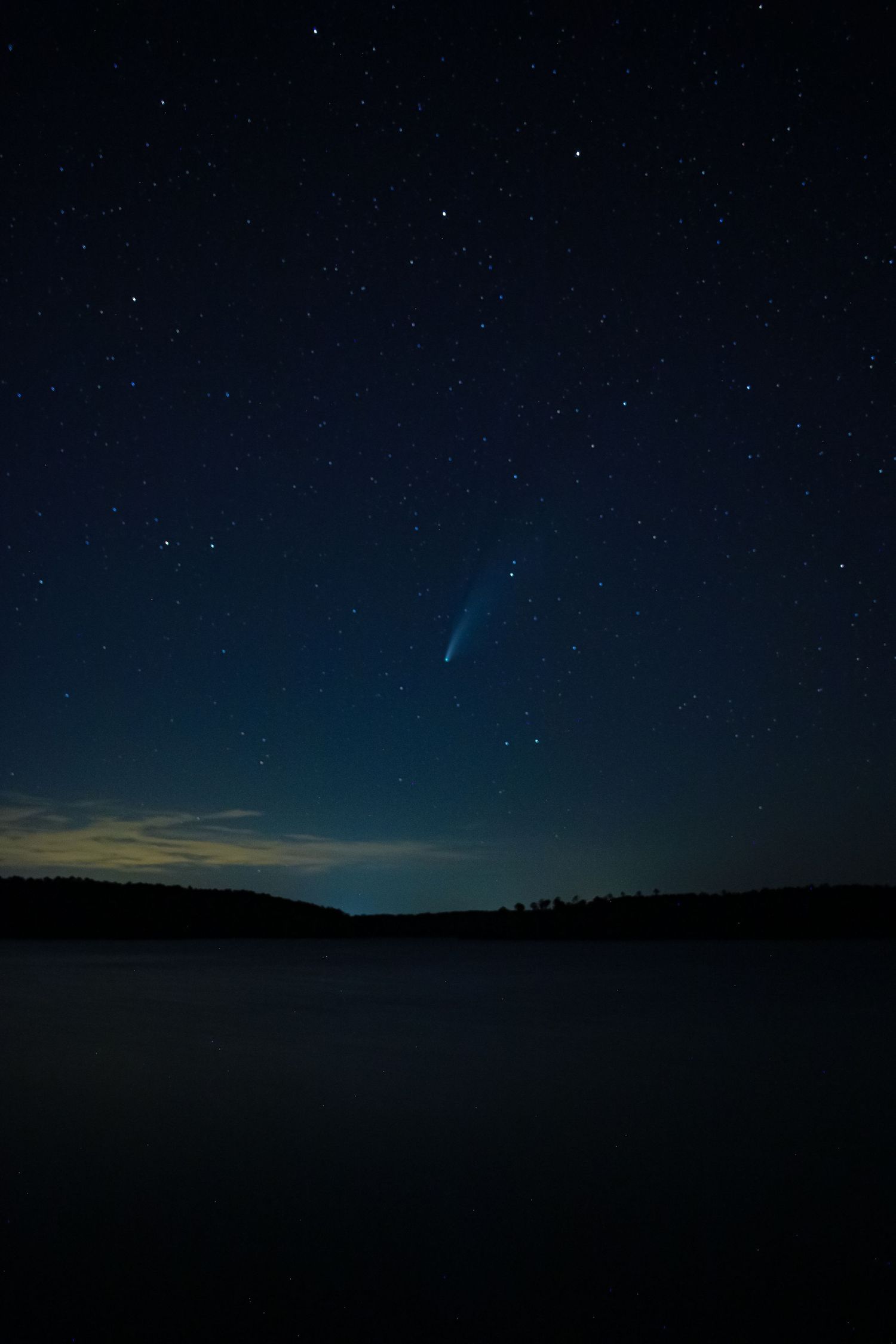 A comet streaks across the starry night sky over a dark landscape silhouette with a faint glow on the horizon. Comet NEOWISE