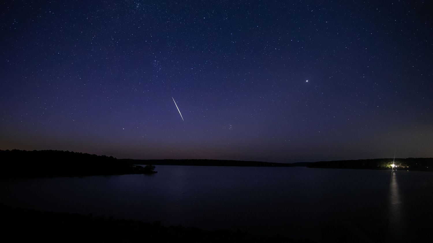 A panoramic view of night sky showing the progression of Starlink sattelite crossing the sky light across the horizon over a lake.