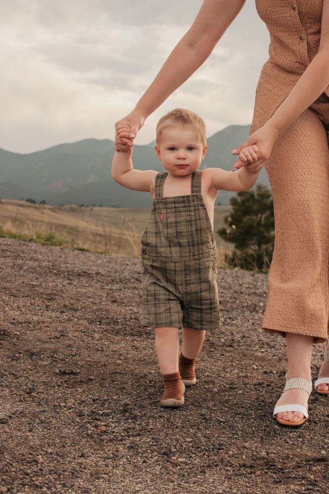 Toddler in plaid overalls learning to walk outdoors held by mother in tan dress with mountains | What to Wear for Family Photos