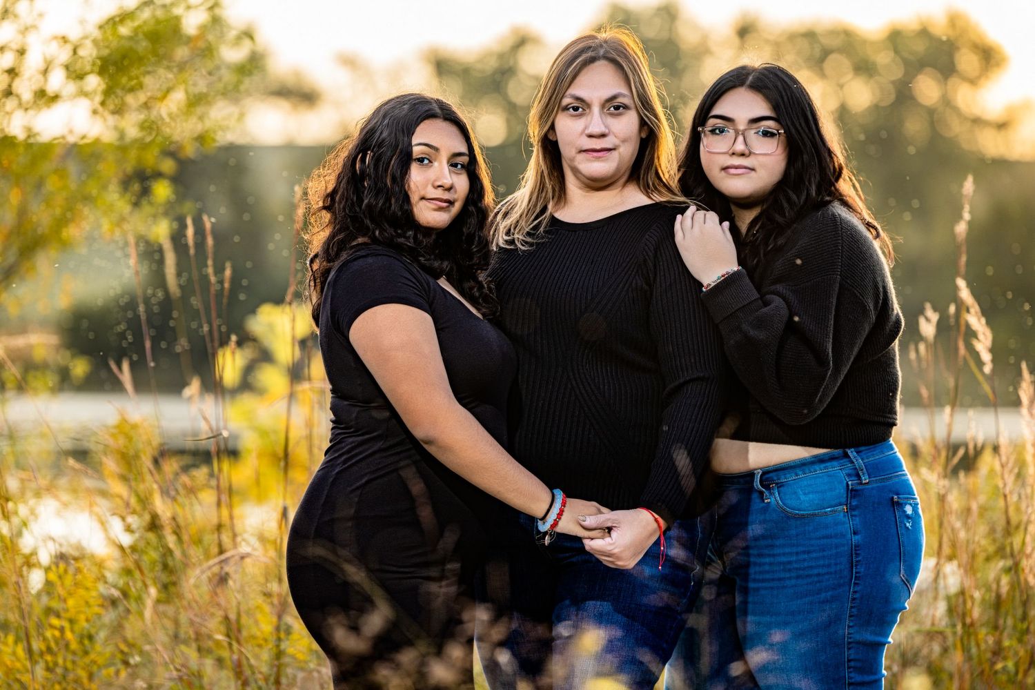 Three young women posing together outdoors at golden hour near a lake in casual black outfits.