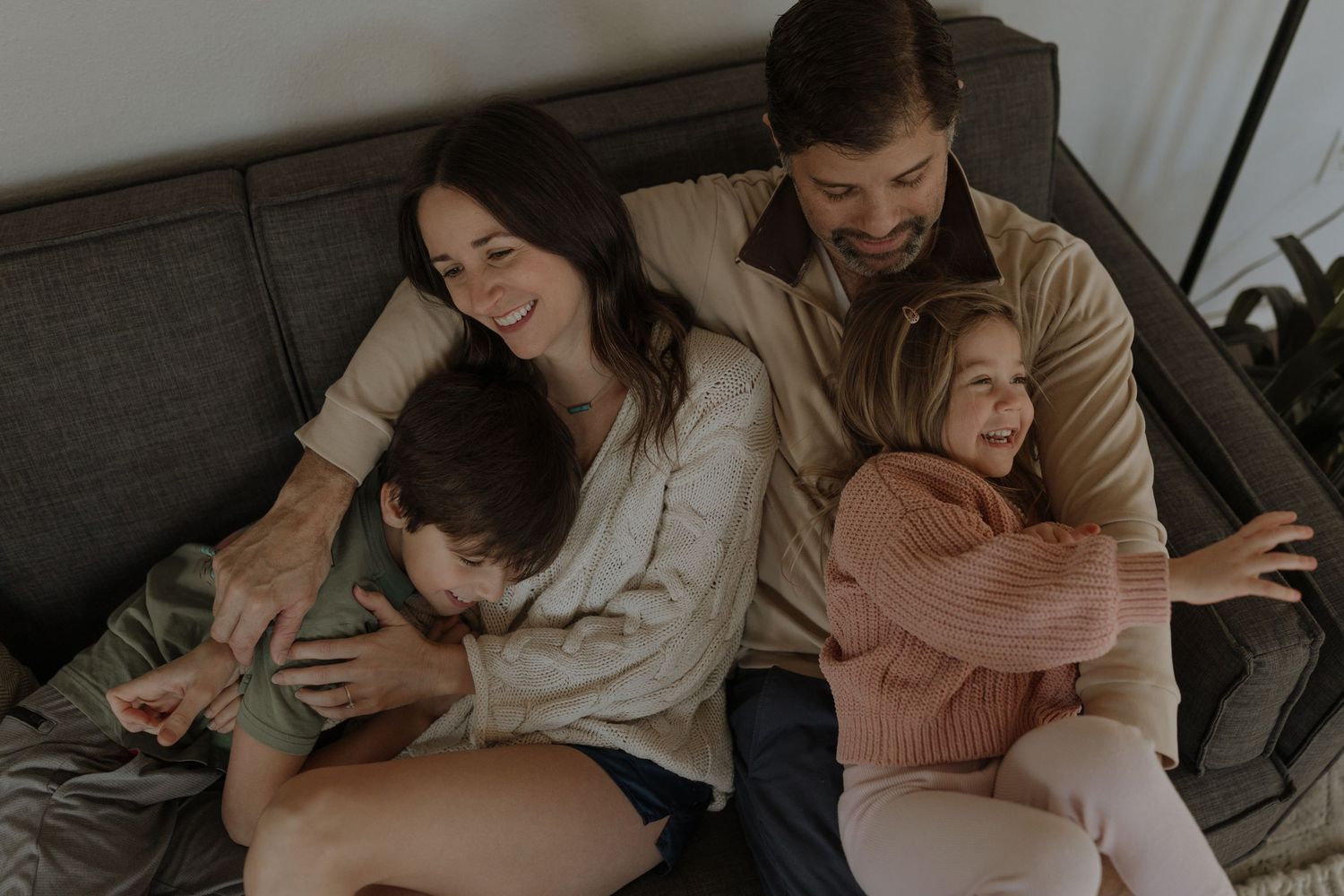 A family snuggles together on a grey couch in a cozy living room setting.