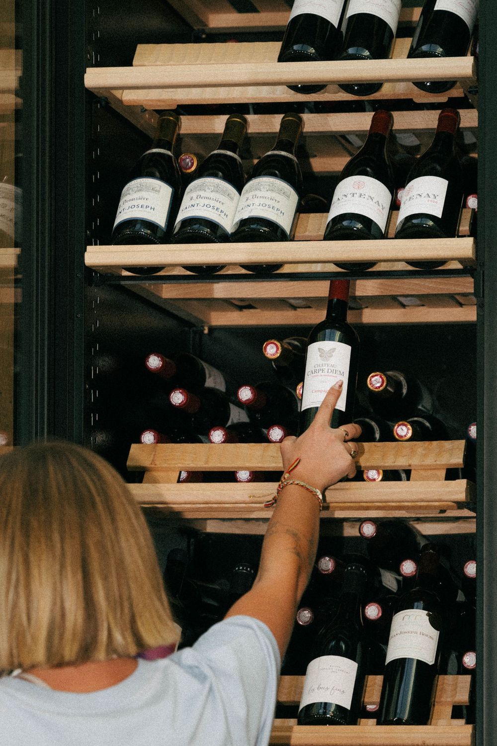 Woman selecting a red wine bottle from a wooden wine rack filled with various bottles in a wine cellar.